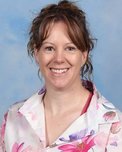 A woman with shoulder-length red hair wearing a light blue patterned top, smiling, against a light blue background.