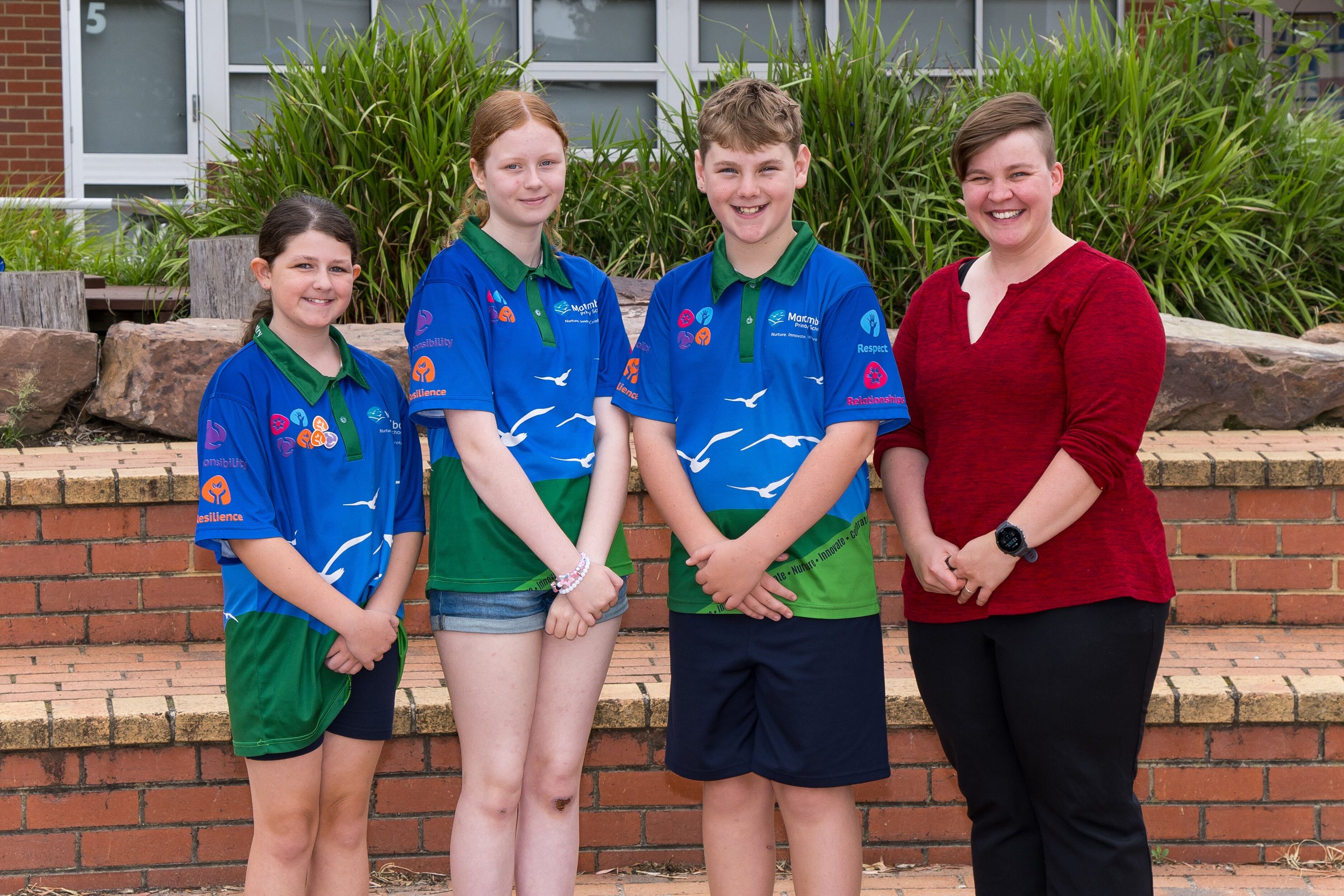 Three children standing outdoors in front of a colorful mural with trees and bushes in the background. Two of them are in blue school uniforms with a snake design, and one is in a navy and aqua shirt.