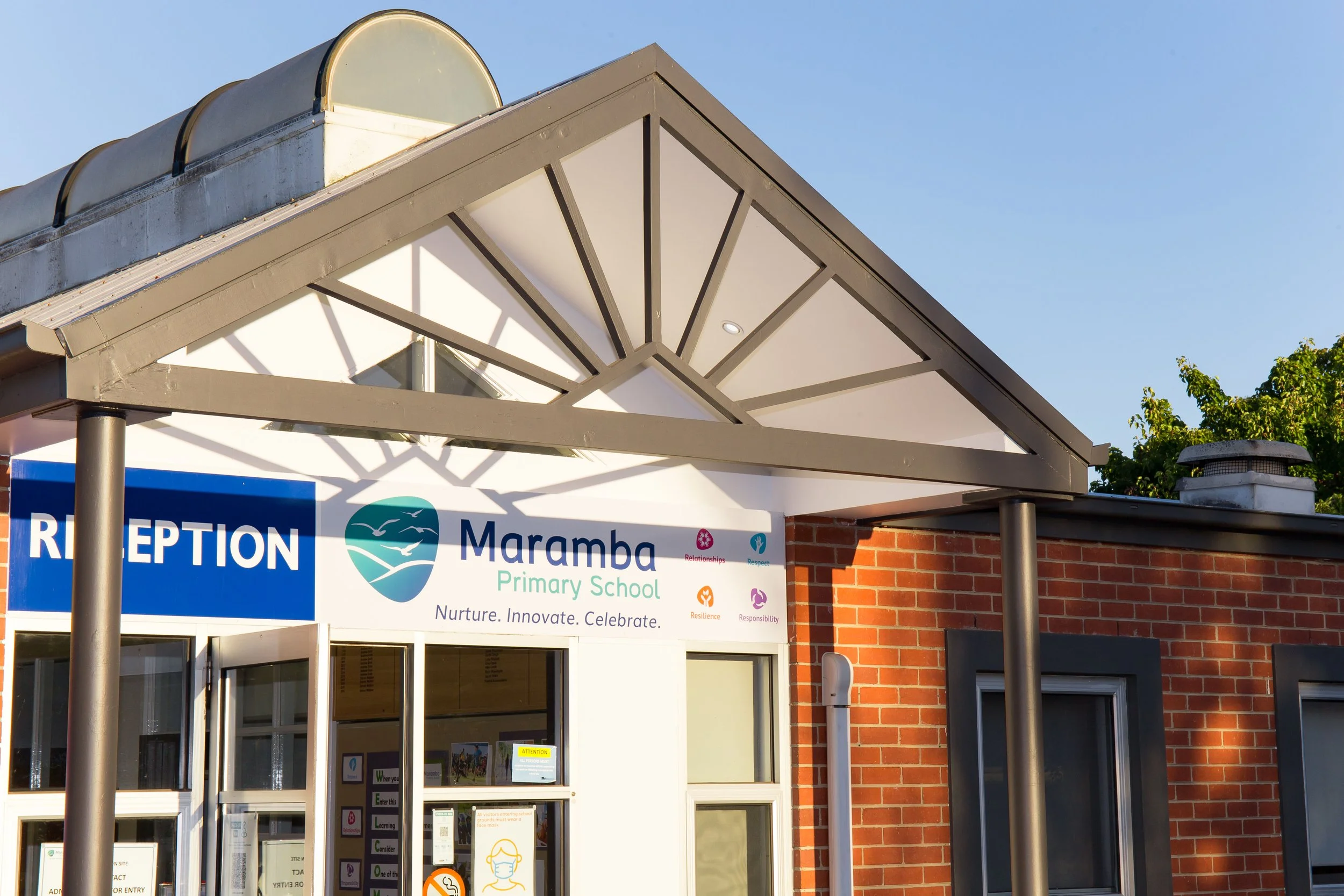 The entrance of Maramba Primary School with a sign displaying the school's name, logo, and mottos, under a metal-framed canopy on a brick building exterior.