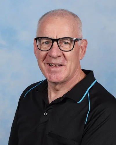 A portrait of an older man with glasses, short gray hair, wearing a striped button-up shirt against a light blue background.