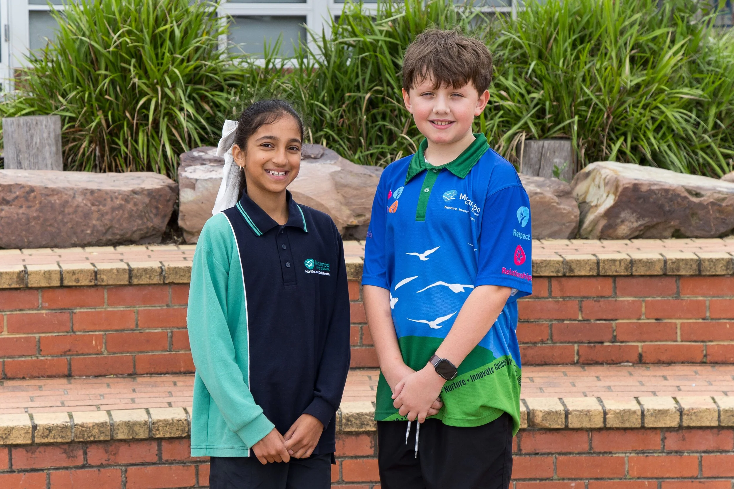 Three students standing outdoors in front of a colorful mural, wearing blue shirts with Australian animals and school logo, smiling at the camera.