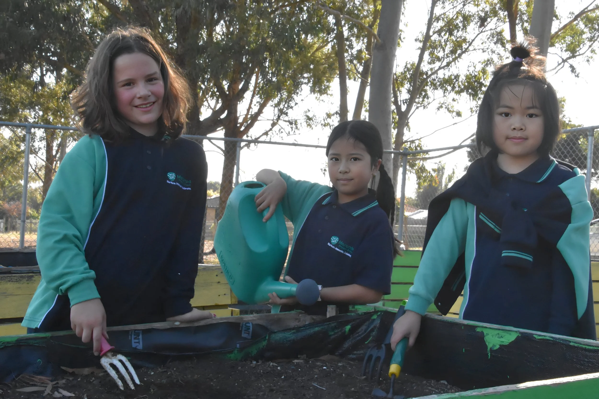 Three young girls in school uniforms gardening outdoors with tools and watering can.