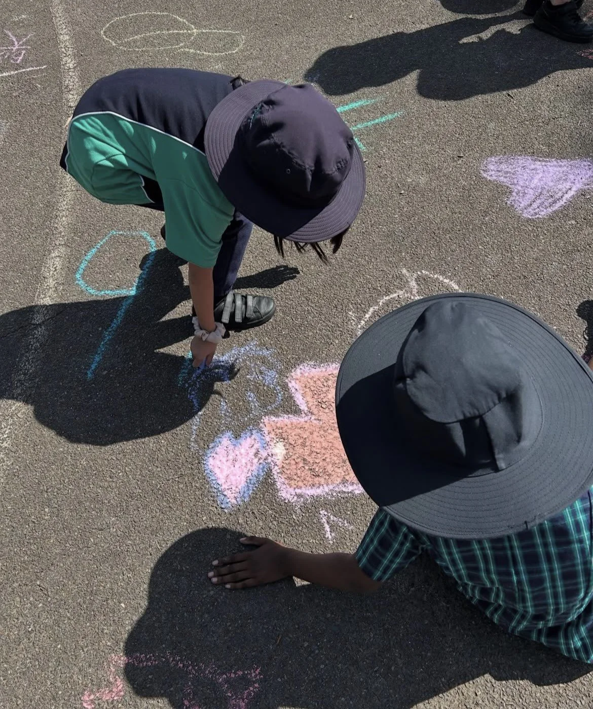 Two children wearing wide-brimmed hats drawing with chalk on pavement, creating heart shapes and other designs.