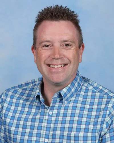 Portrait of a smiling man with short spiky hair, wearing a light gray button-up shirt, against a blue background.