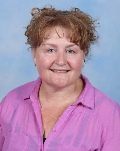 A smiling woman with curly, reddish-blonde hair wearing a black top and a gold necklace with a pendant, against a light blue background.