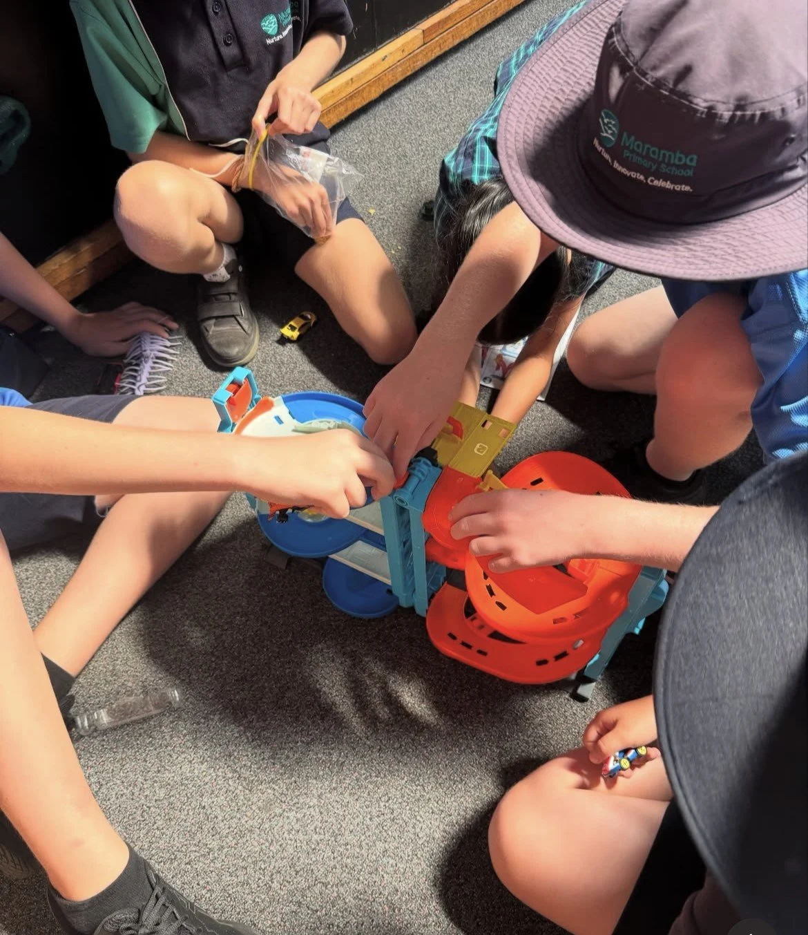 A group of children playing a marble run game on carpeted floor, with some children sitting and others kneeling, wearing casual clothing and hats.