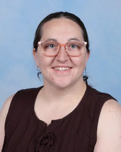 A woman with long brown hair, wearing glasses and a dark blue dress with white polka dots, smiling in front of a light blue background.