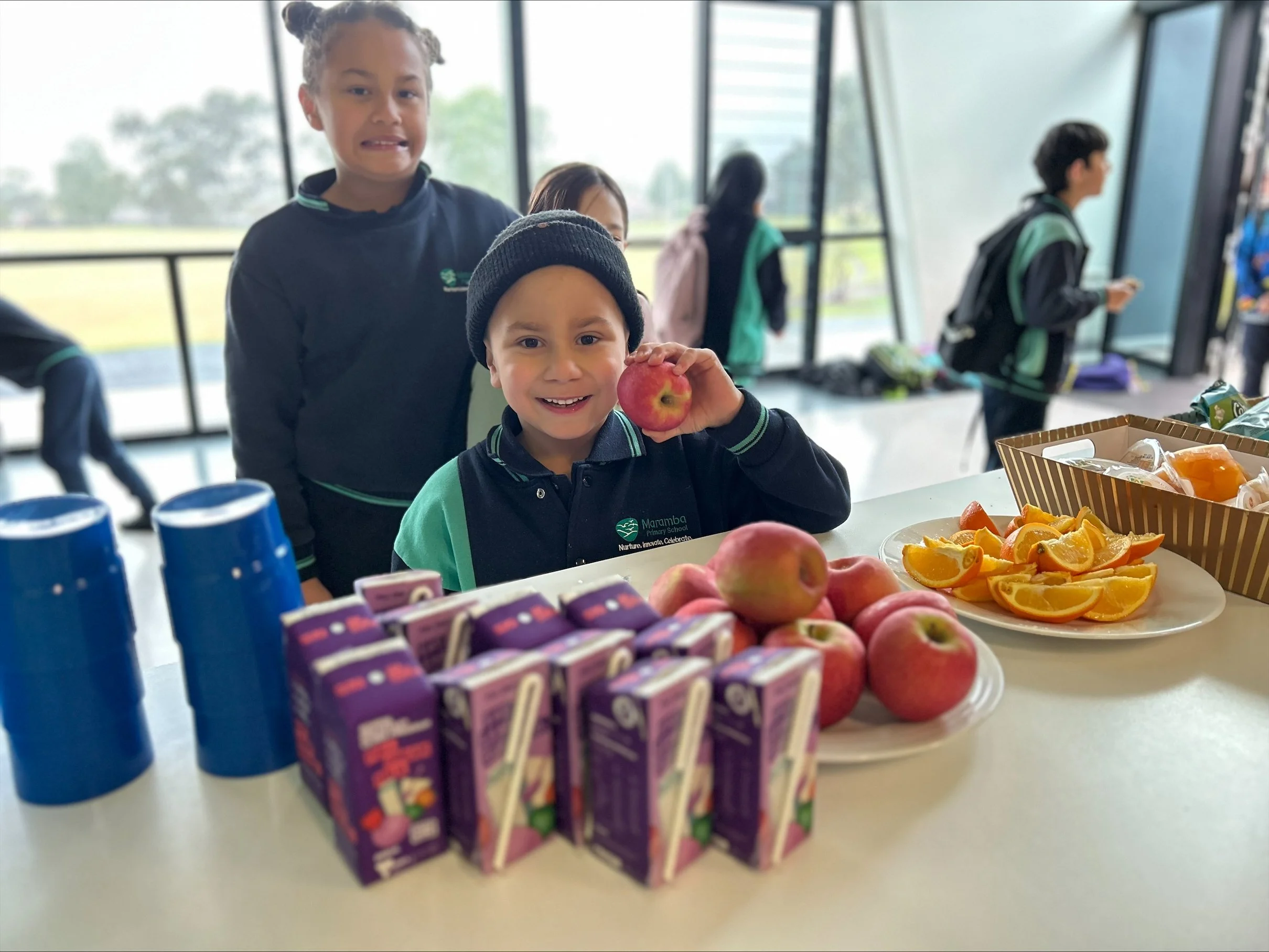 Two children, a girl and a boy, standing behind a table filled with apples, orange slices, juice boxes, and cups, inside a bright room with large windows and other children in the background.