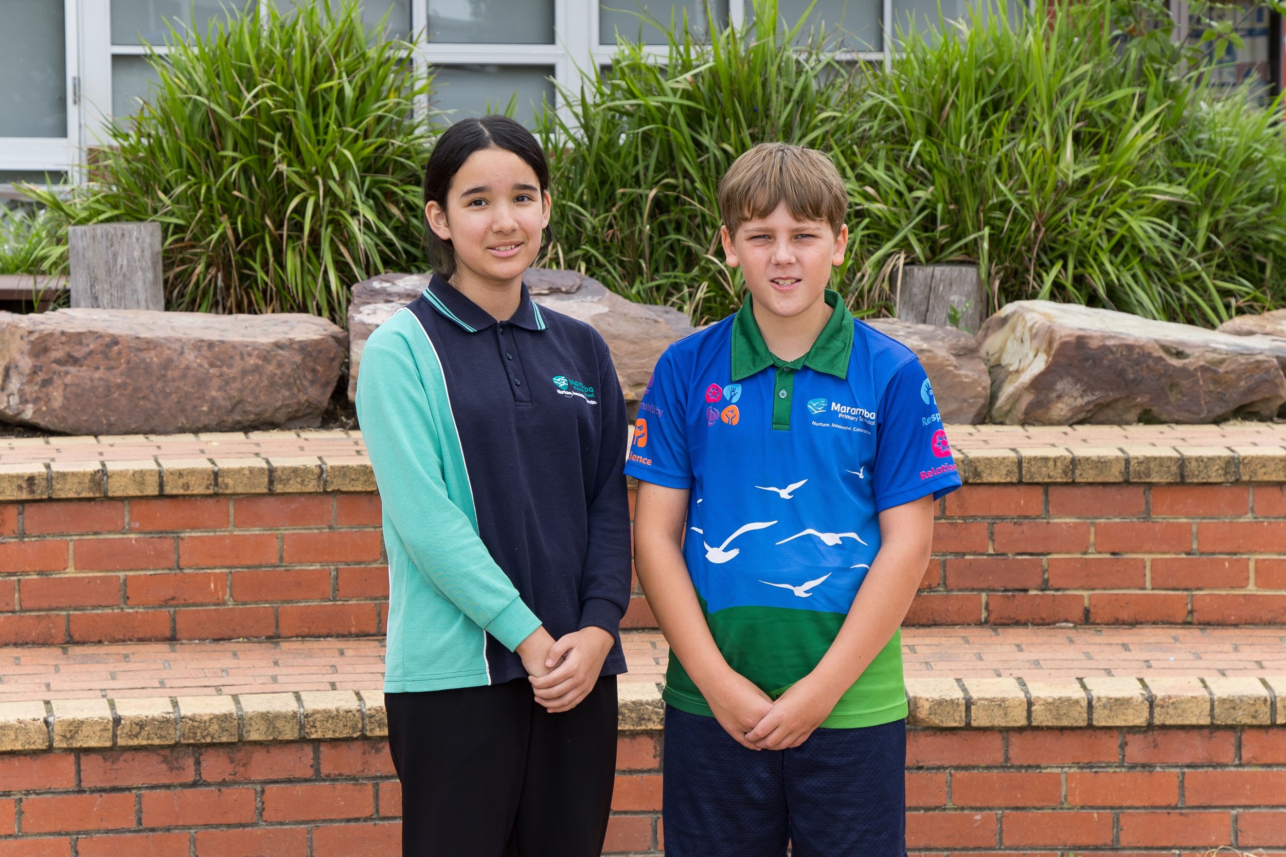 Three students wearing blue shirts with a colorful snake design, standing outdoors in front of a painted mural and greenery.