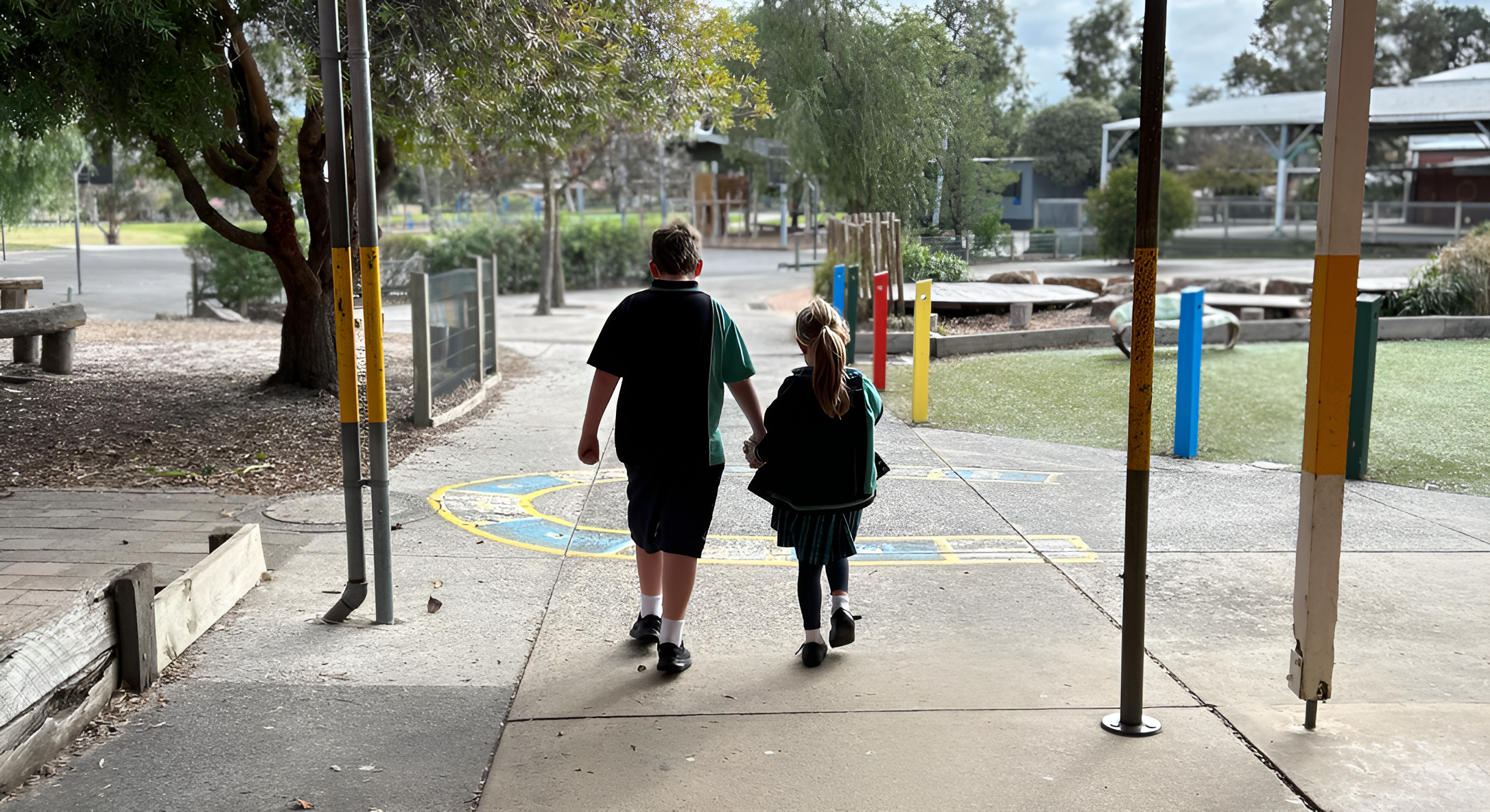 Two children, a boy and a girl, walking hand in hand into a park playground area.