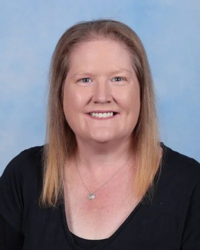 Portrait of a smiling woman with red hair, glasses, and a black top with ruffled sleeves against a blue background.