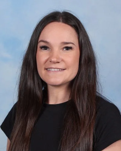Portrait of a woman with long dark hair, wearing a black top, smiling slightly against a light blue background.