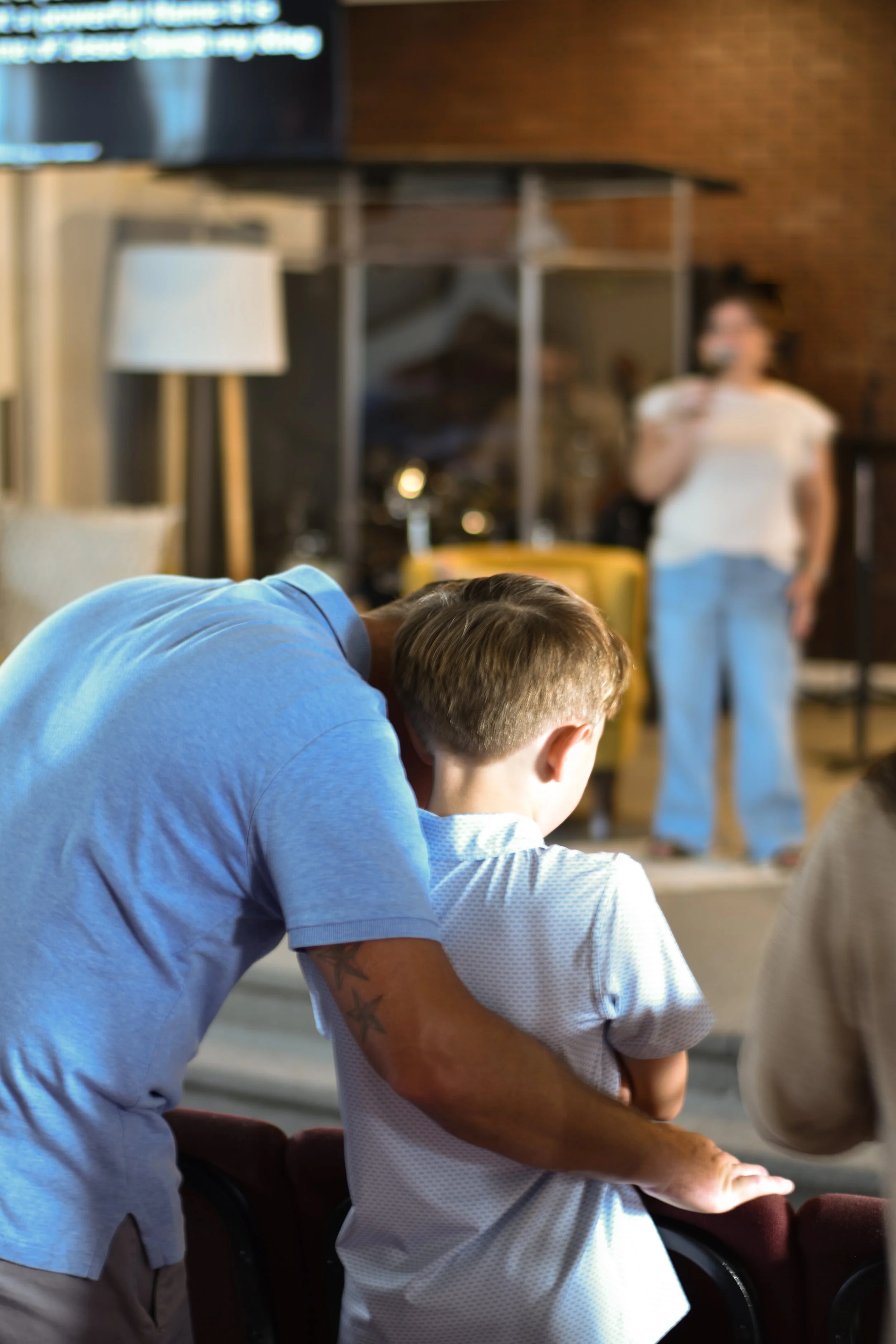 A father leaning over his young son in a blue shirt, speaking to him, in a room with a woman in the background holding a microphone during praise and worship.