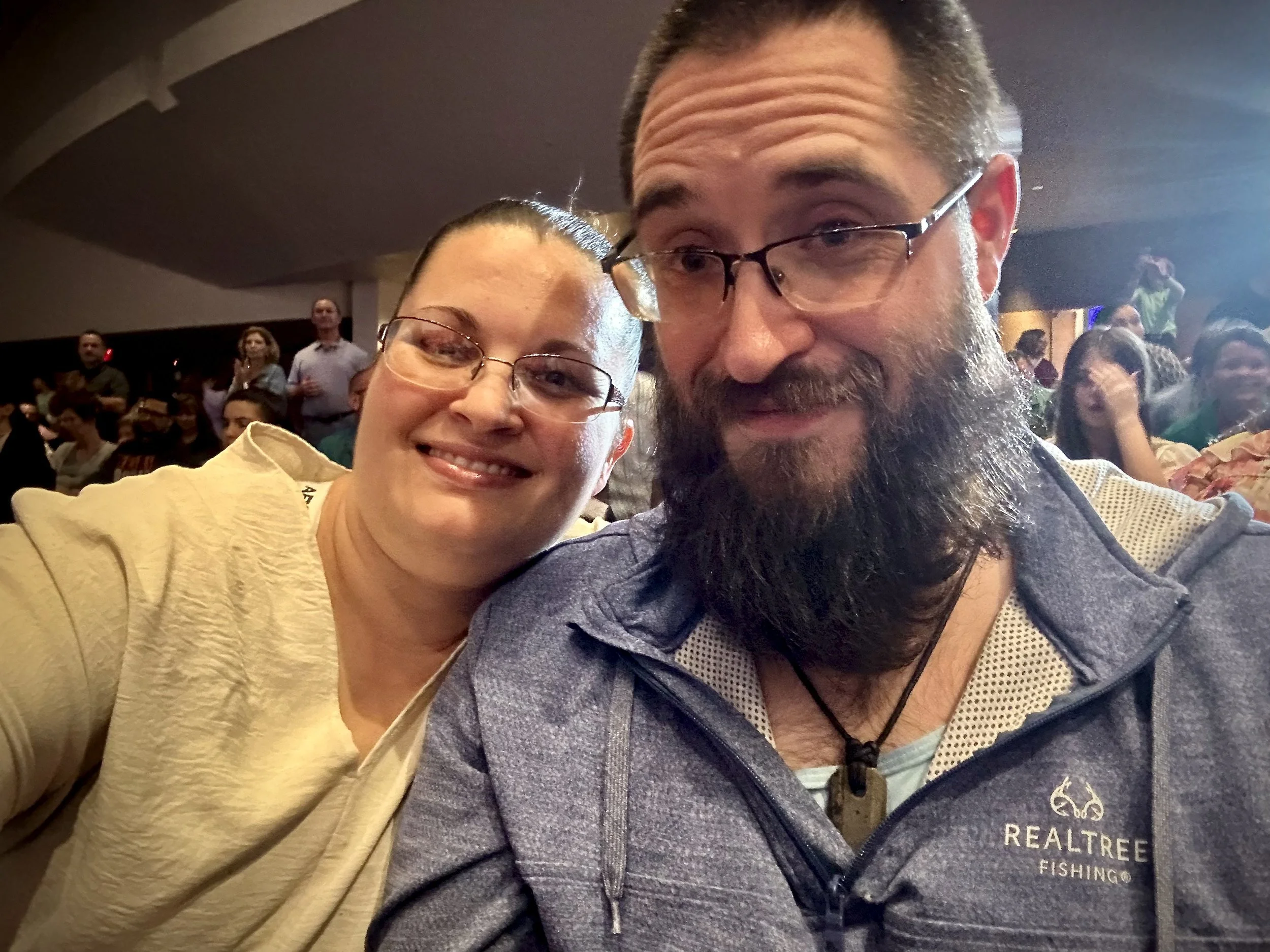 A smiling woman and a bearded man taking a selfie at an indoor event with multiple people in the background.