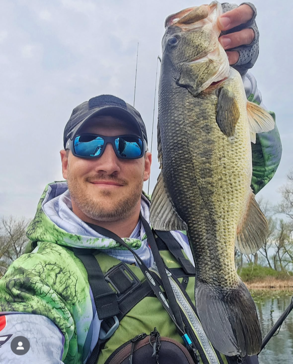 Man wearing sunglasses and outdoor gear holding a large fish he caught, with a fishing rod in the background, near a body of water and trees.