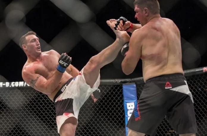 Two male mixed martial artists compete in a UFC fight inside the octagon, with one delivering a high kick to the other's face.