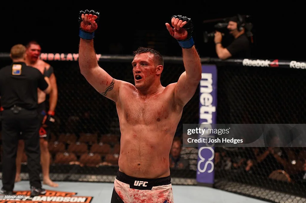 A victorious male mixed martial artist in the UFC, shirtless and wearing gloves, raising his arms in triumph inside the octagon.
