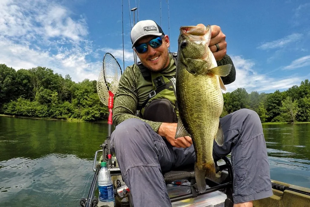 Man in fishing gear sitting in a boat on a lake, holding a large bass fish.