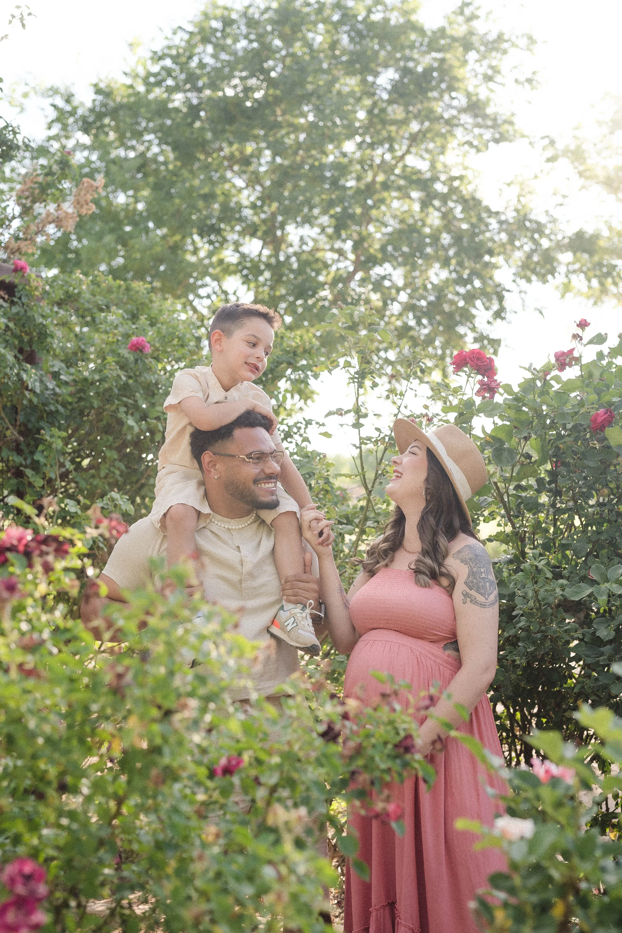 A joyful family shares a playful moment in a lush garden, with a young boy riding on his father's shoulders with the mother, smiling and pregnant, stands beside them - captured by Paul Porras in Albuquerque, New Mexico