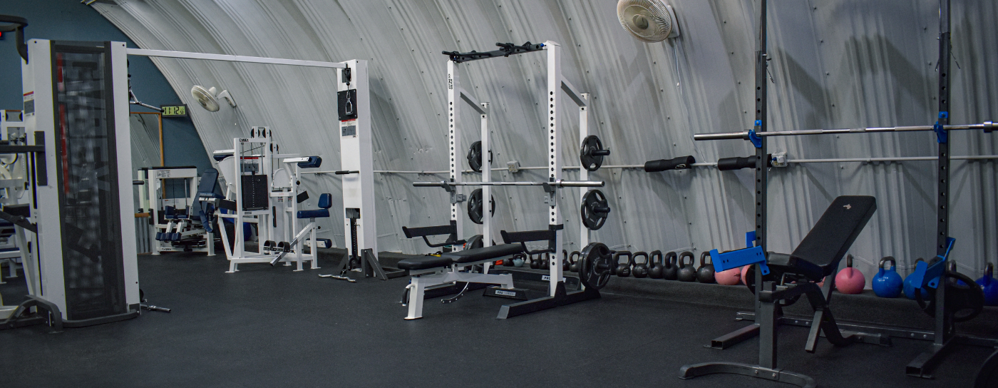 Interior of a gym with various workout equipment including a bench, weight plates, kettlebells, and an exercise machine, all set against a curved, gray wall.