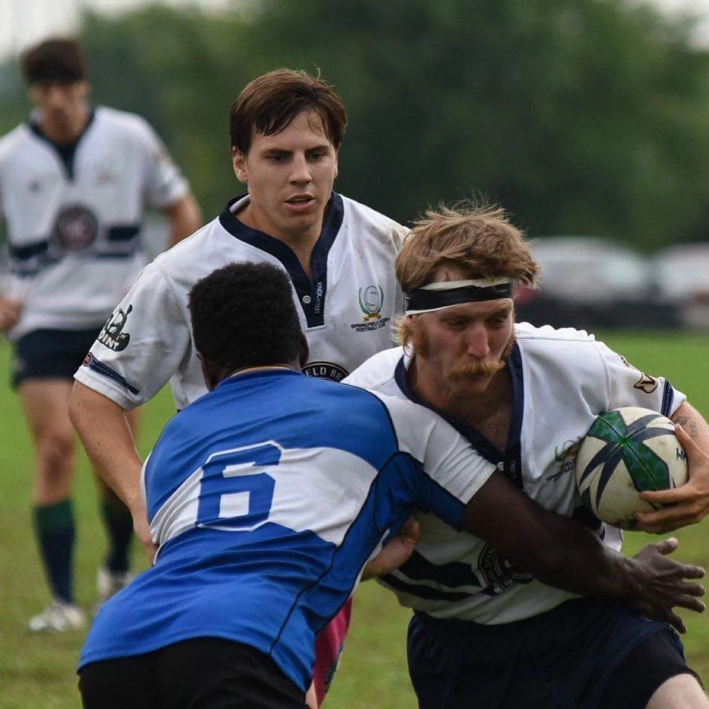 A rugby match in progress with one player holding the ball while being tackled by another player, and two teammates approaching from behind on a grassy field.