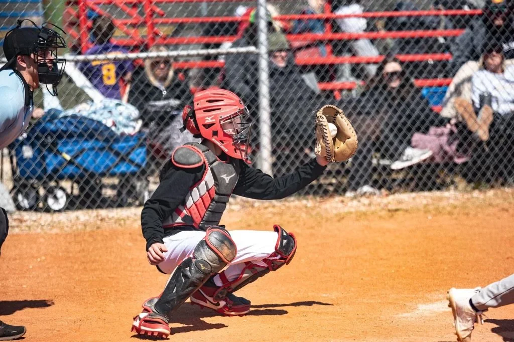 Young baseball catcher in black and red gear squatting on orange dirt, catching a pitch, with spectators behind a chain-link fence.