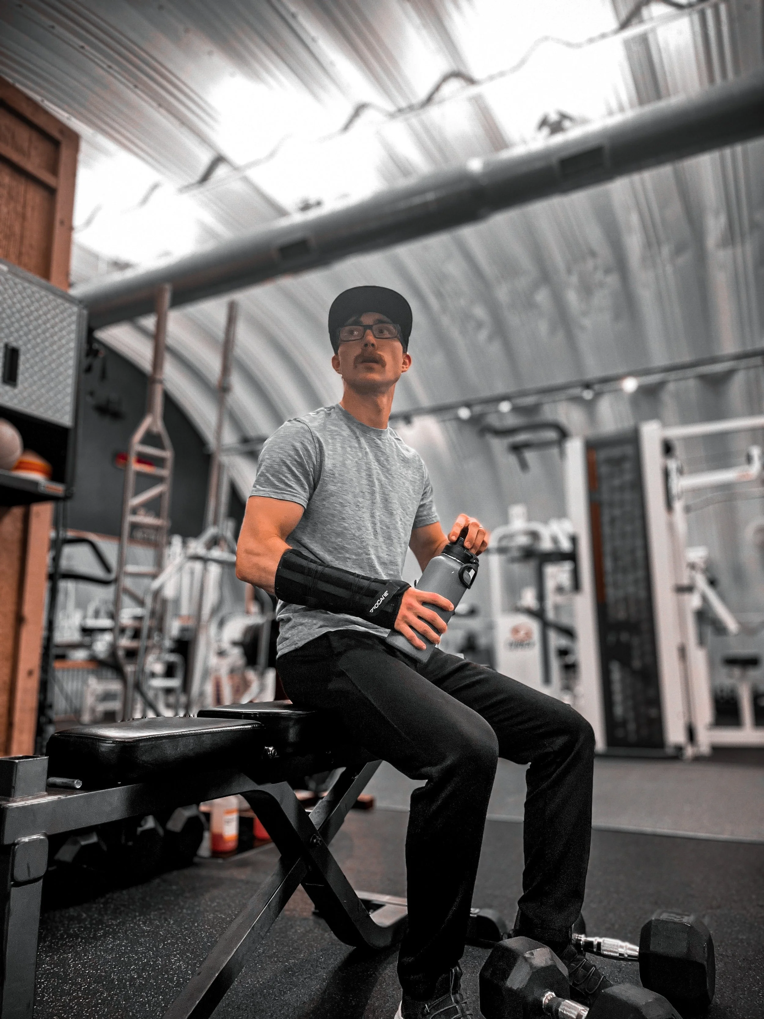 A man sitting on a workout bench in a gym, holding a water bottle, wearing a gray t-shirt, black pants, a black cap, glasses, and a black arm brace. There is gym equipment in the background.