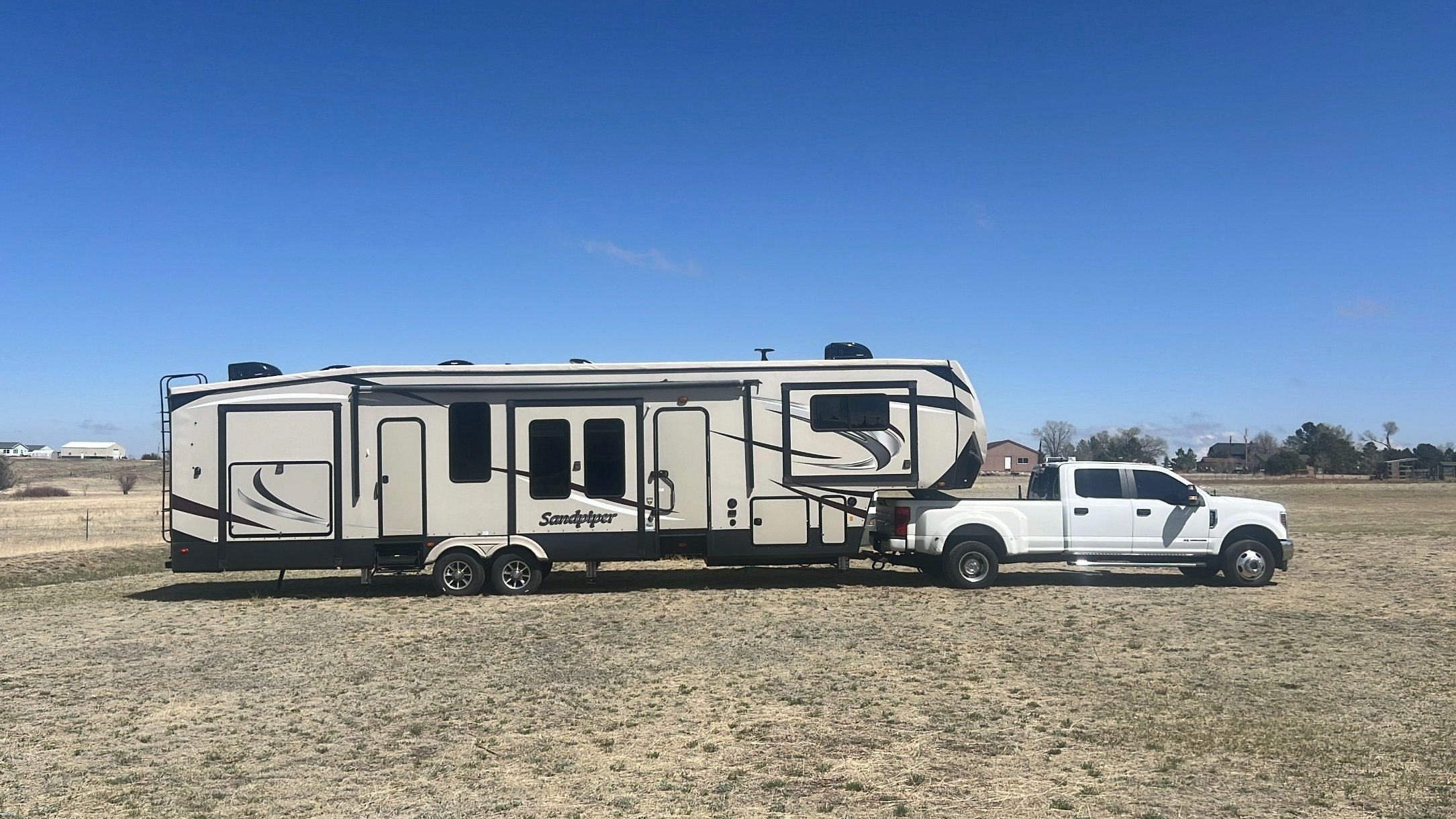 A white pickup truck attached to a large beige and black Sandpiper travel trailer parked on a grassy field under a clear blue sky.