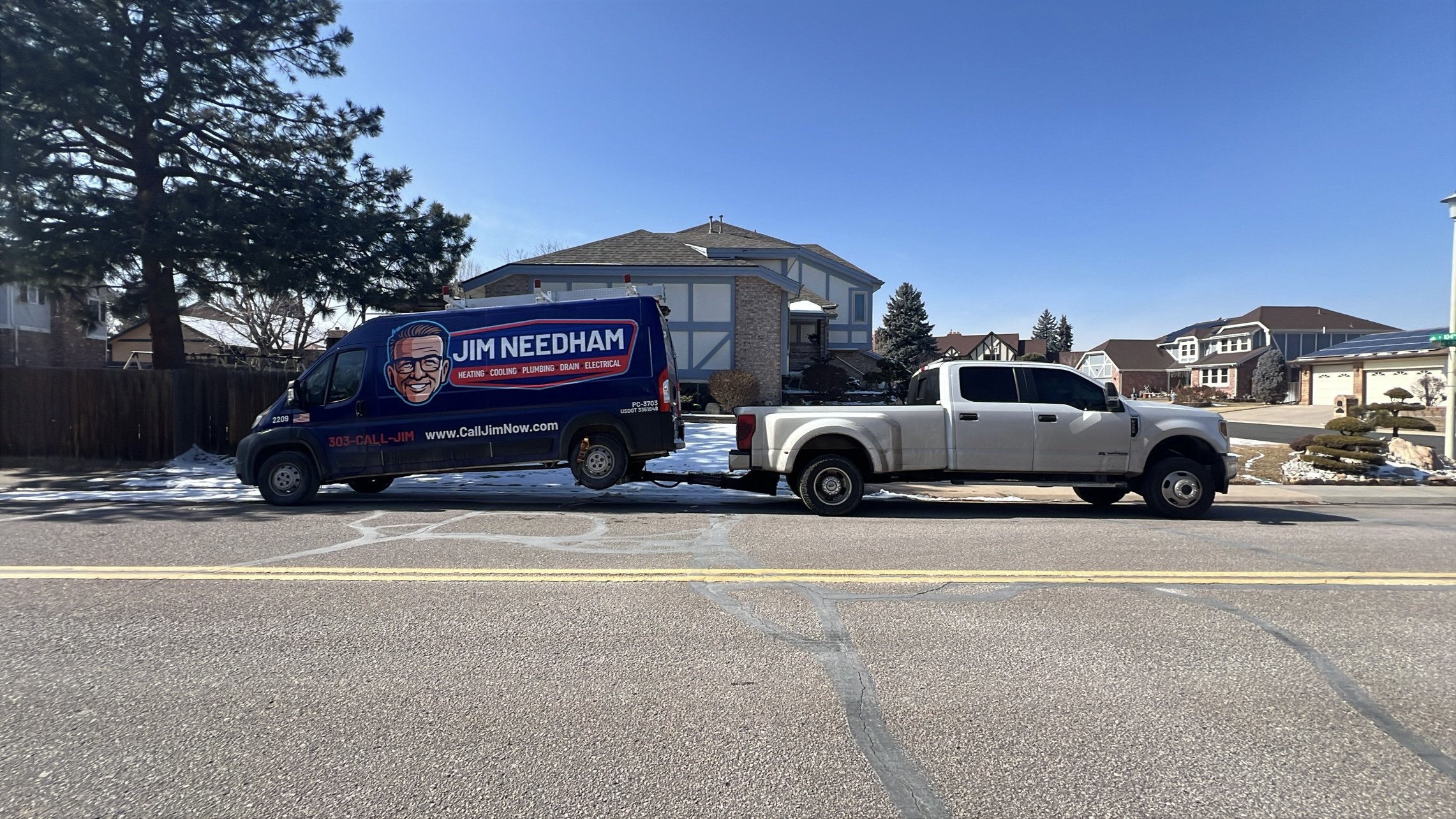 A blue Jim Needham service van being towed by a white pickup truck on a residential street with houses and a tree in the background.