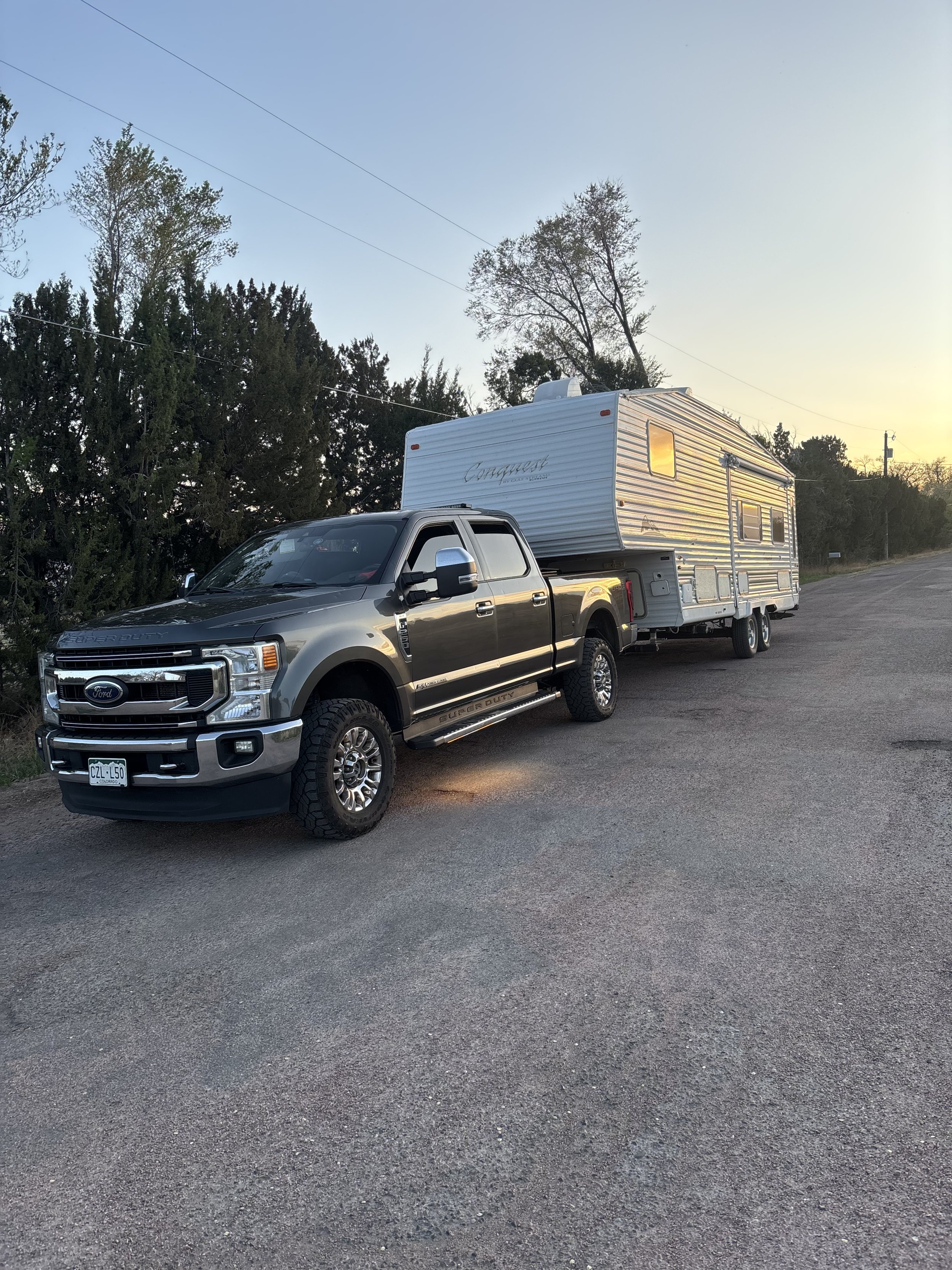 A Ford pickup truck towing a travel trailer parked on a gravel roadside during sunset.