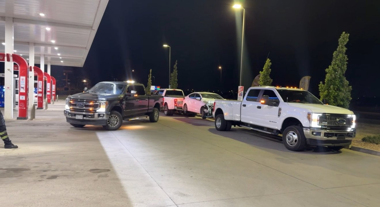 Nighttime scene at a gas station with multiple vehicles, including a white pickup truck parked near the pumps, and two damaged cars in the background, one with front-end damage.