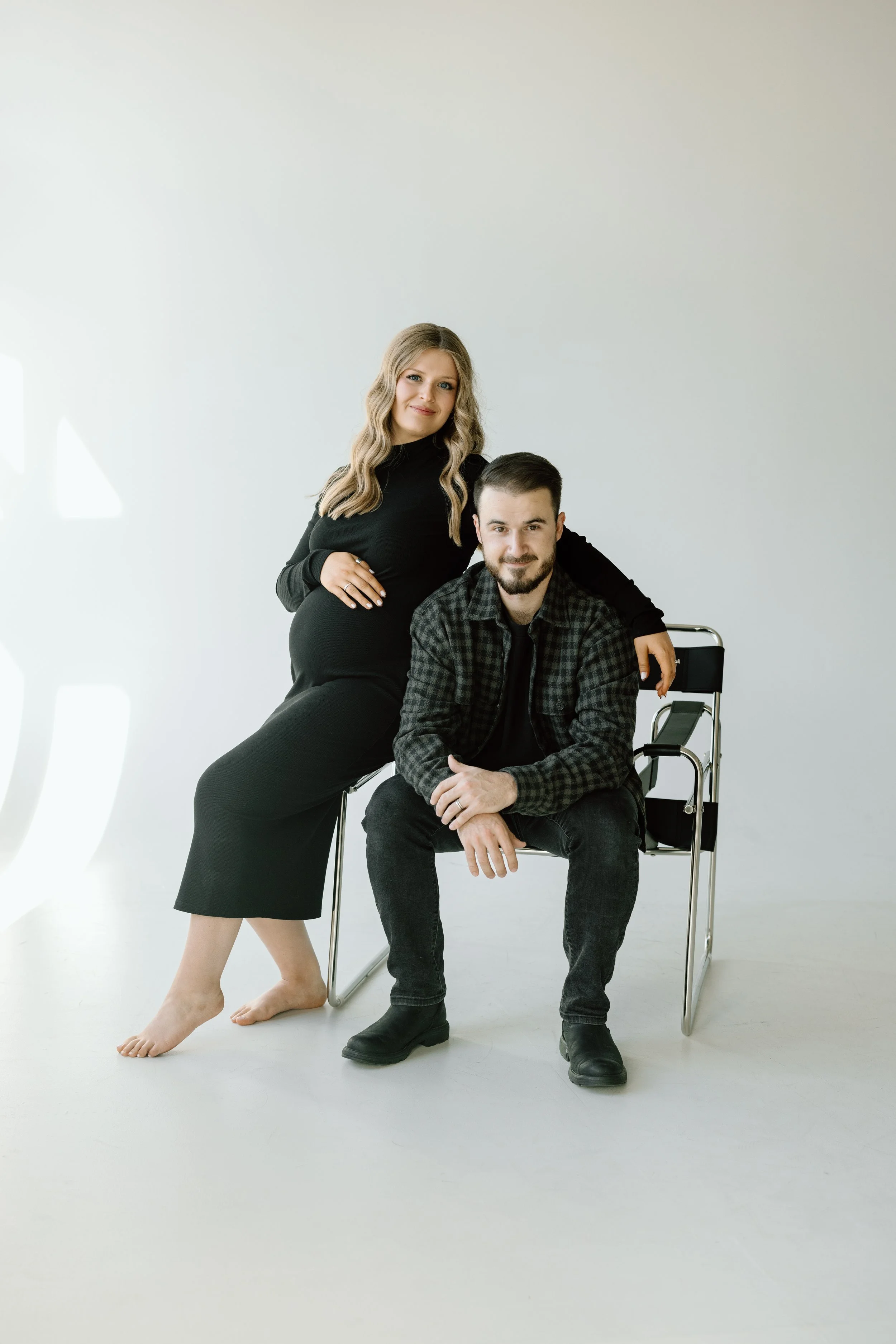 A pregnant woman in a black dress standing beside a seated man in a black and gray plaid shirt, both in a minimalist studio setting.