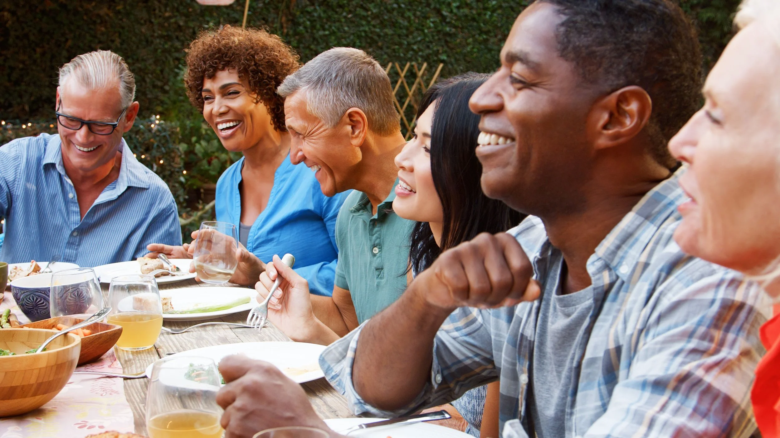 A group of people smiling and laughing at a dinner party outdoors with food and drinks on a wooden table.