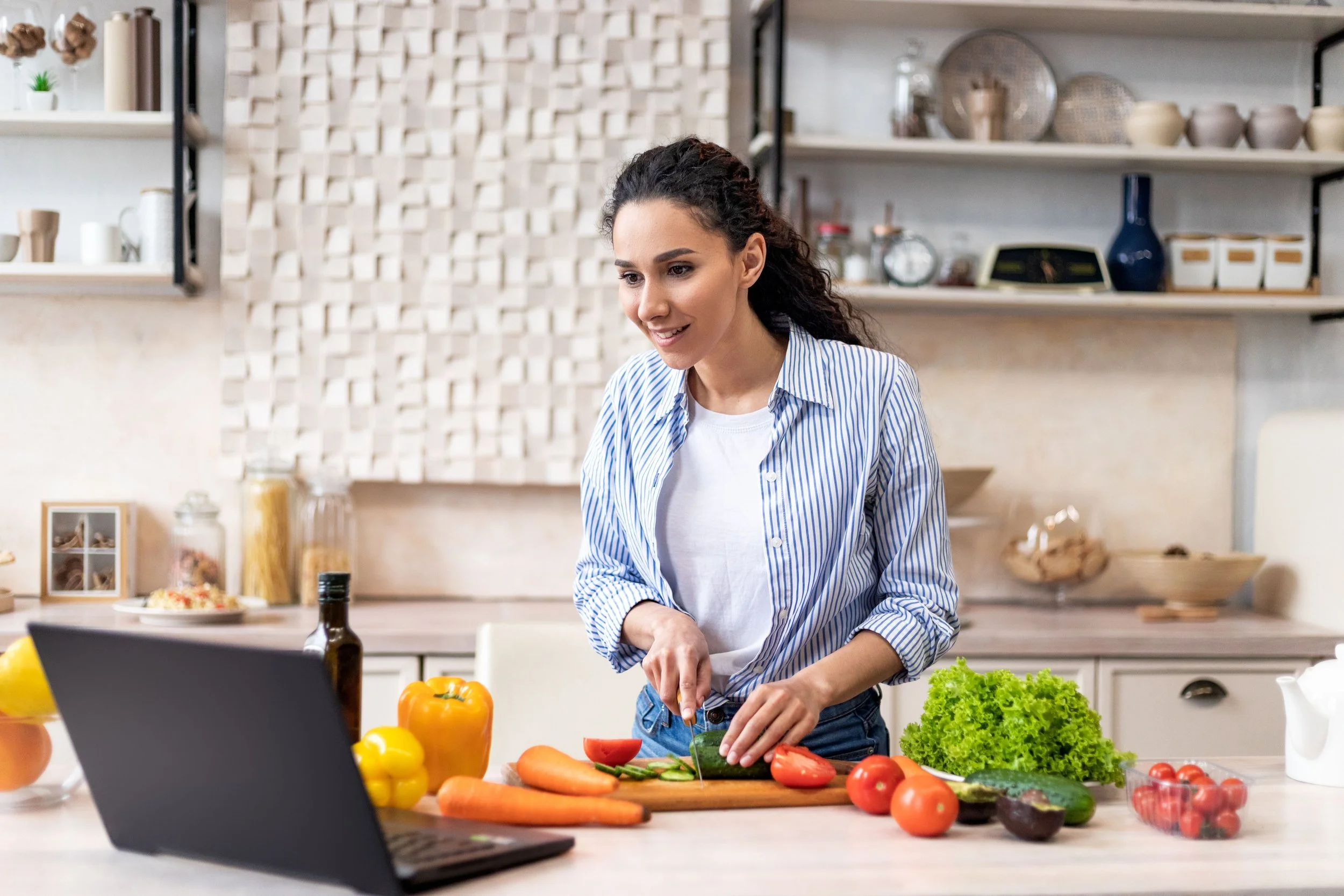 Woman in a kitchen chopping vegetables for a recipe, with fresh produce on the counter and a laptop in front of her.
