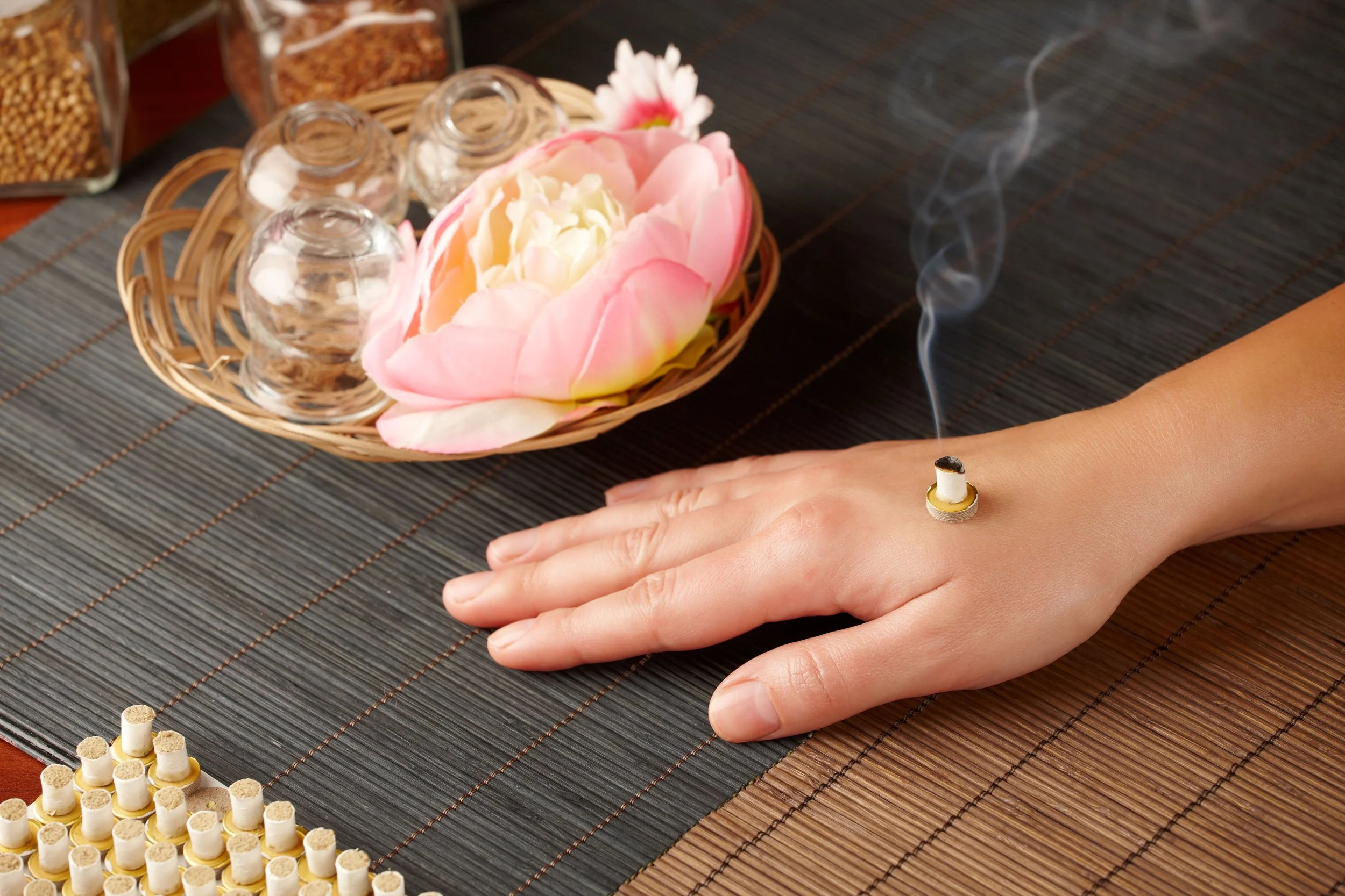A person receiving an incense massage, with a lit incense stick on their hand, on a woven mat with a pink flower, glass bowls, and herbal jars nearby.