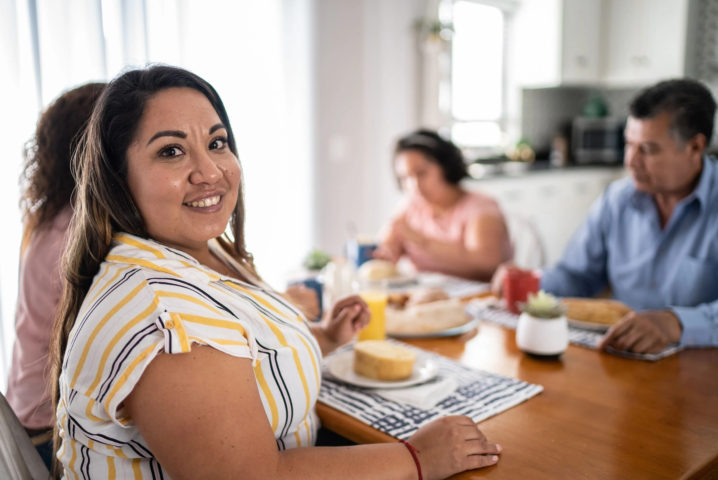 A woman smiling at the camera during a breakfast gathering with two men and one woman in the background, seated at a wooden table with food and drinks.