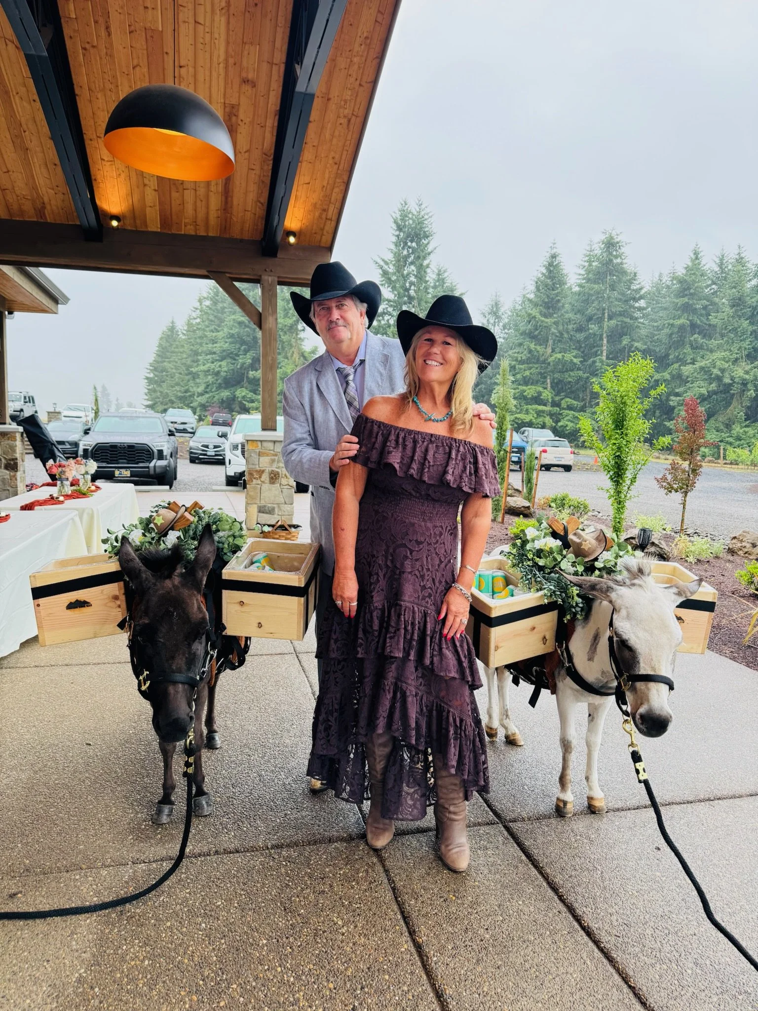 A man and woman wearing cowboy hats and dressed in western-style clothing standing outdoors under a wooden-covered area, each with a small donkey carrying a wooden box filled with decorative items, with cars and trees in the background on a foggy day
