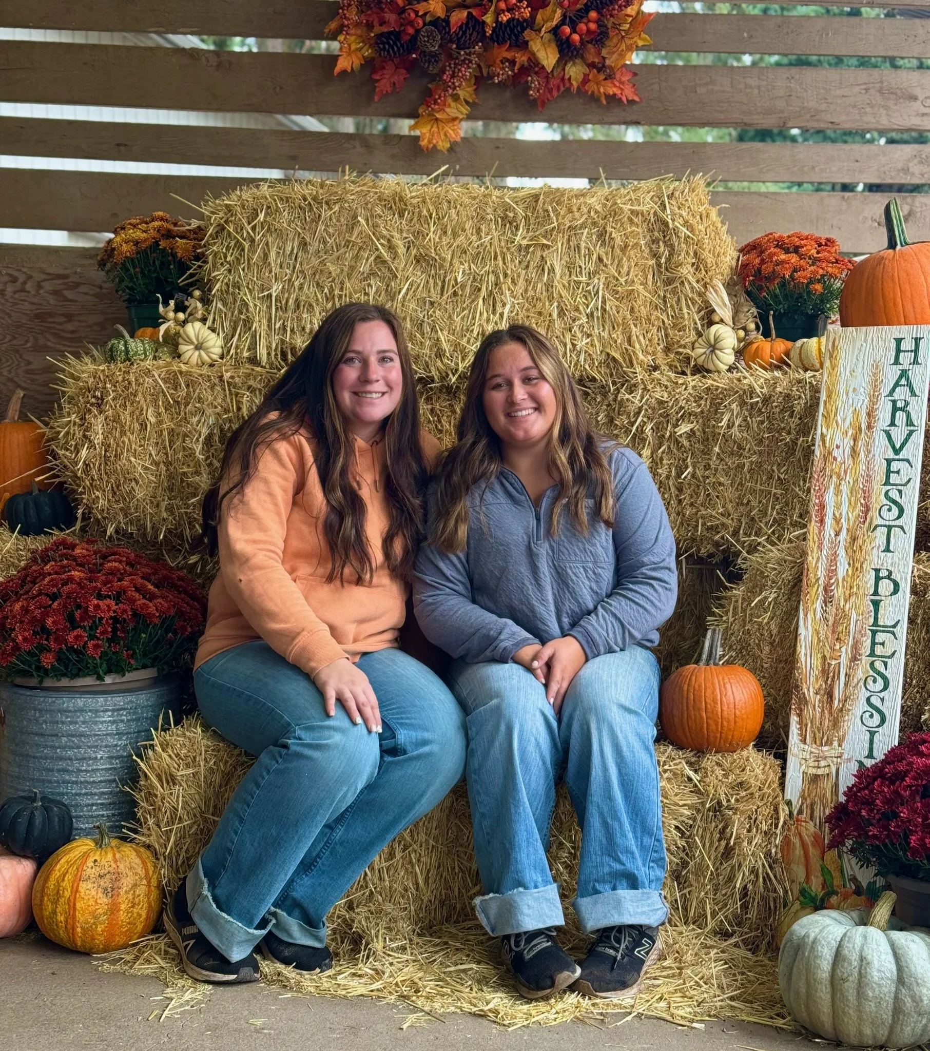 Two women sitting on a hay bale at an autumn festival, surrounded by pumpkins, gourds, and flowers, with a decorative sign that reads 'Harvest Blessings'.