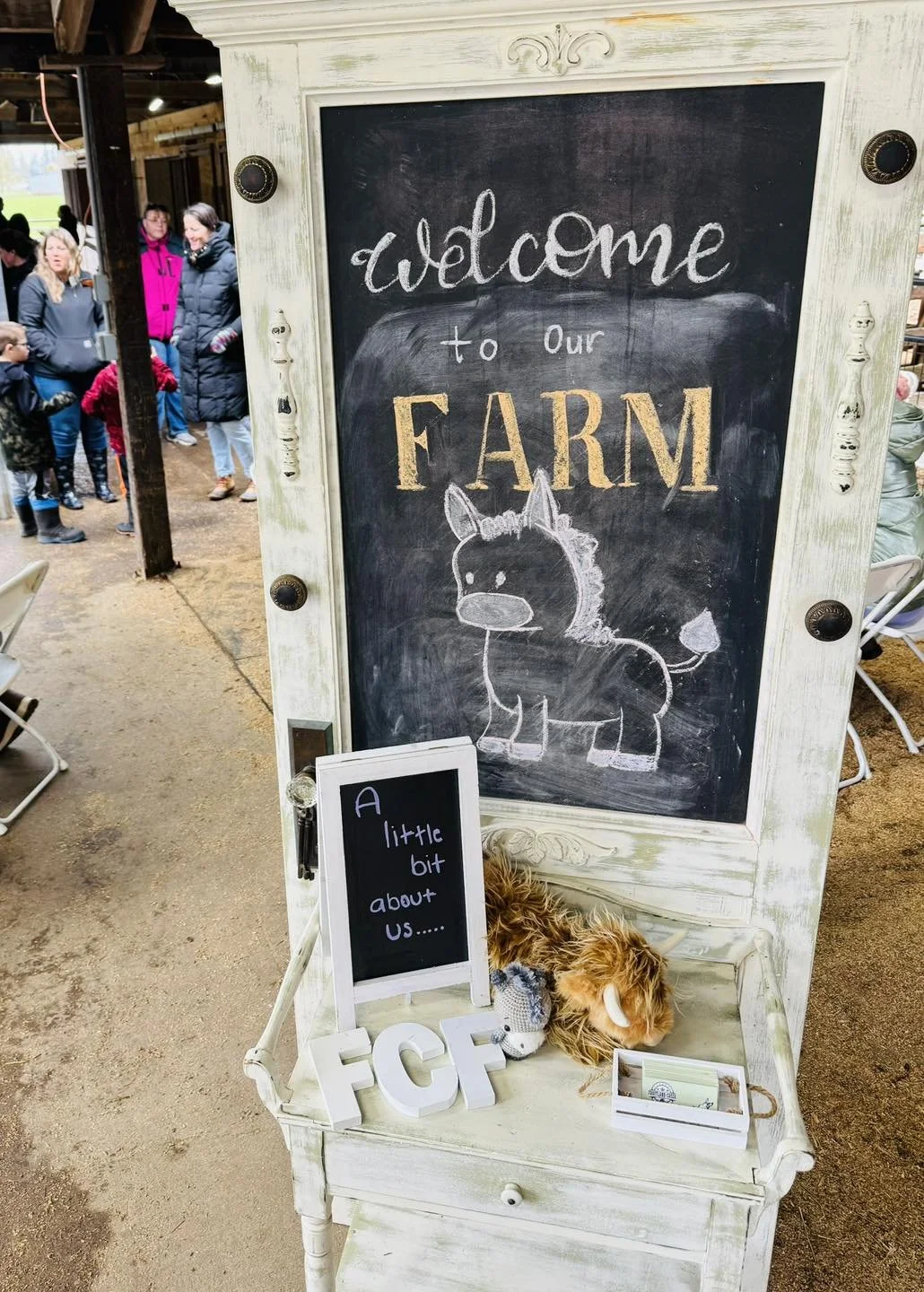 A welcome sign at a farm with a chalkboard message, a chalk drawing of a horse, and plush farm animal toys on a white wooden bench, at an indoor farm attraction.