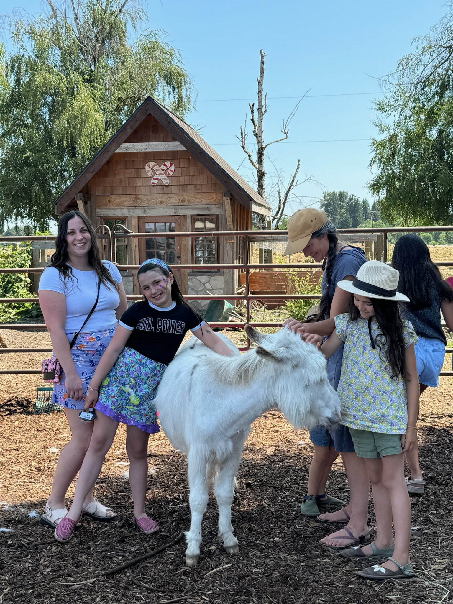 Group of children and a woman interacting with a white goat at a petting farm or zoo, with a wooden barn in the background and trees around.