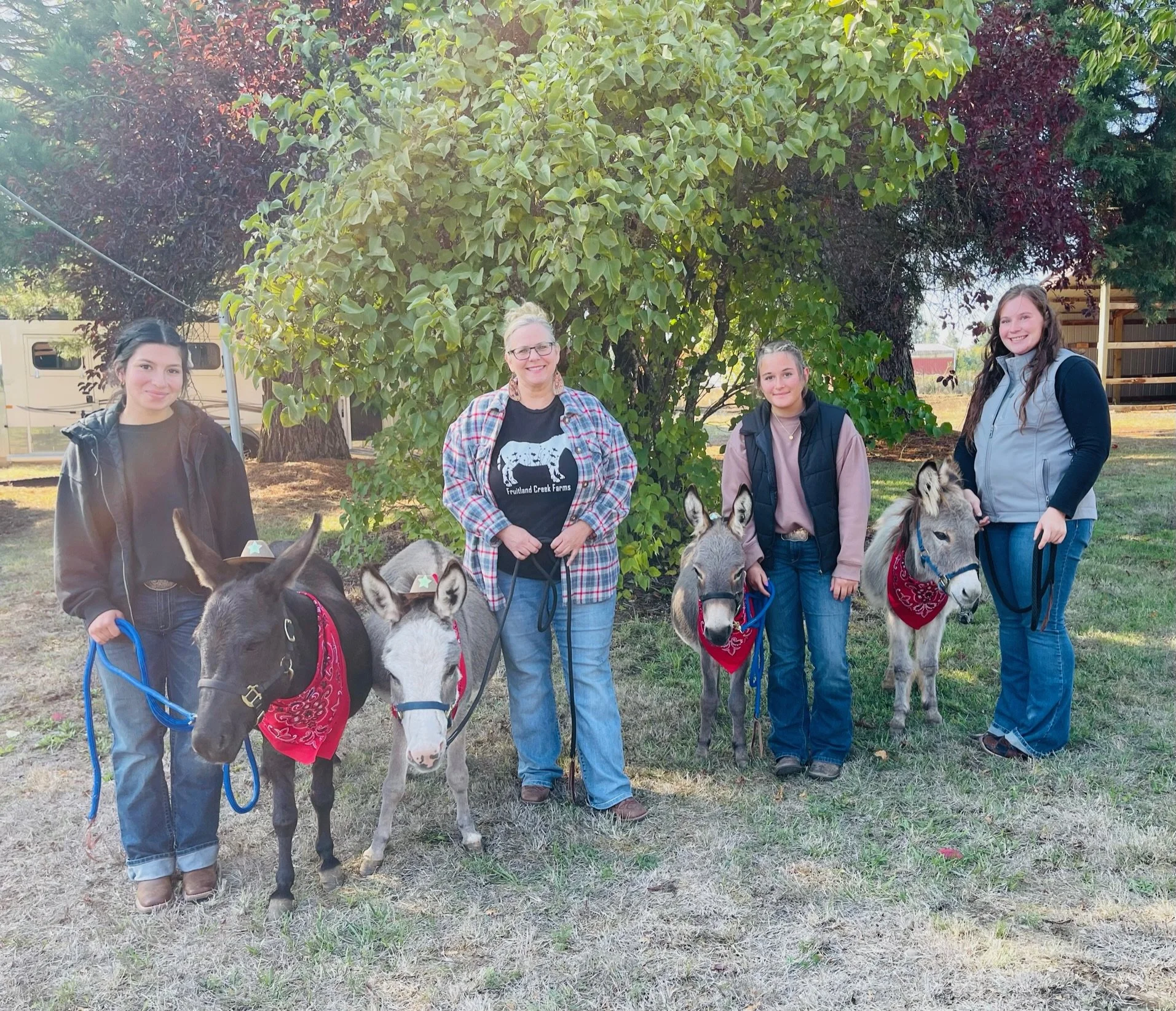 Four women standing outdoors with four donkeys, each woman holding a donkey's lead rope. The women are smiling and standing on a grassy area near a tree, with a vehicle and some fencing visible in the background.