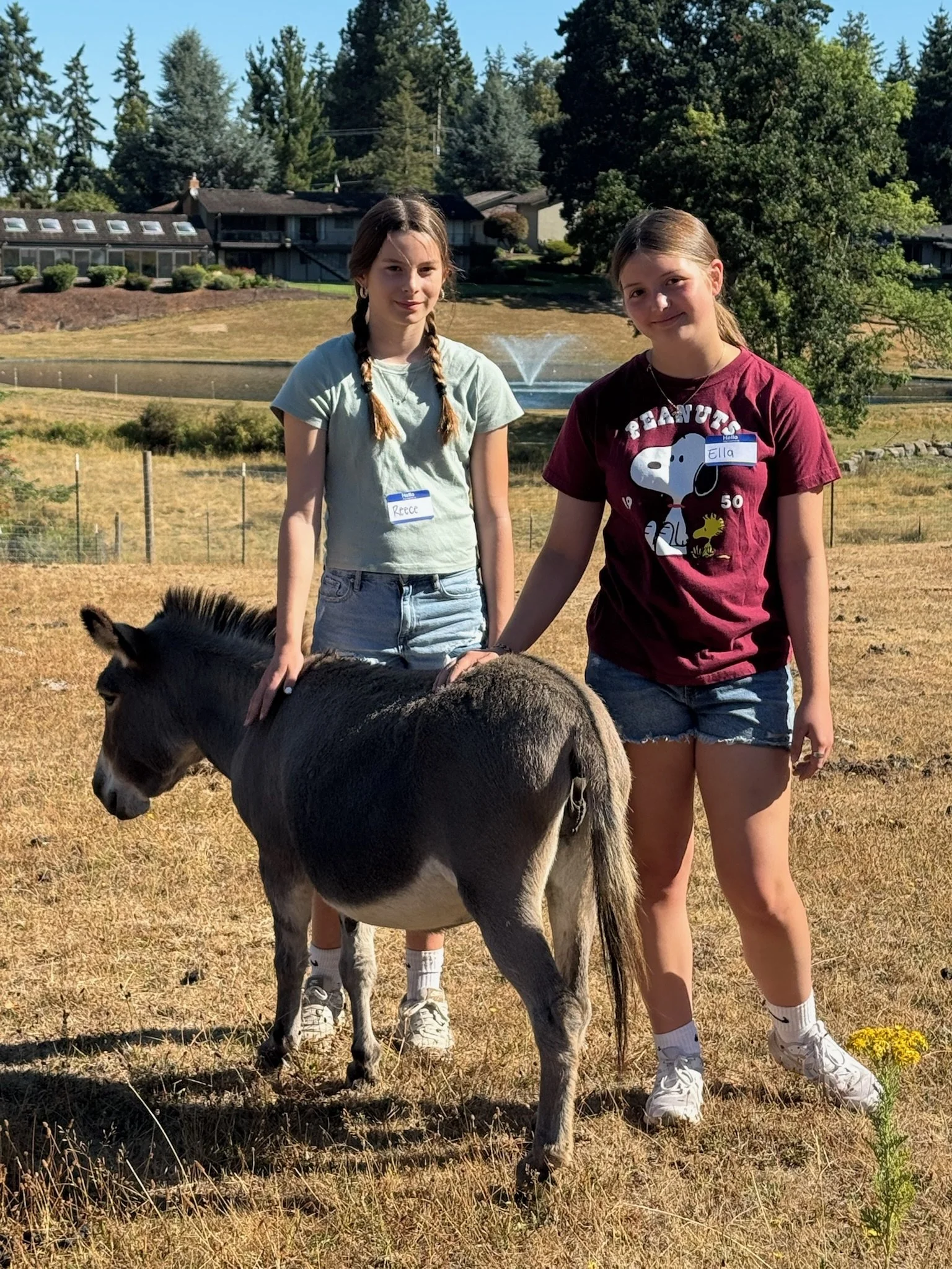 Two young girls holding hands with a donkey in an open field. One girl has braided hair and wears a gray t-shirt, the other has straight hair and wears a maroon Snoopy t-shirt. Name tags reading 'Reece' and 'Ella' are visible. In the background, ther