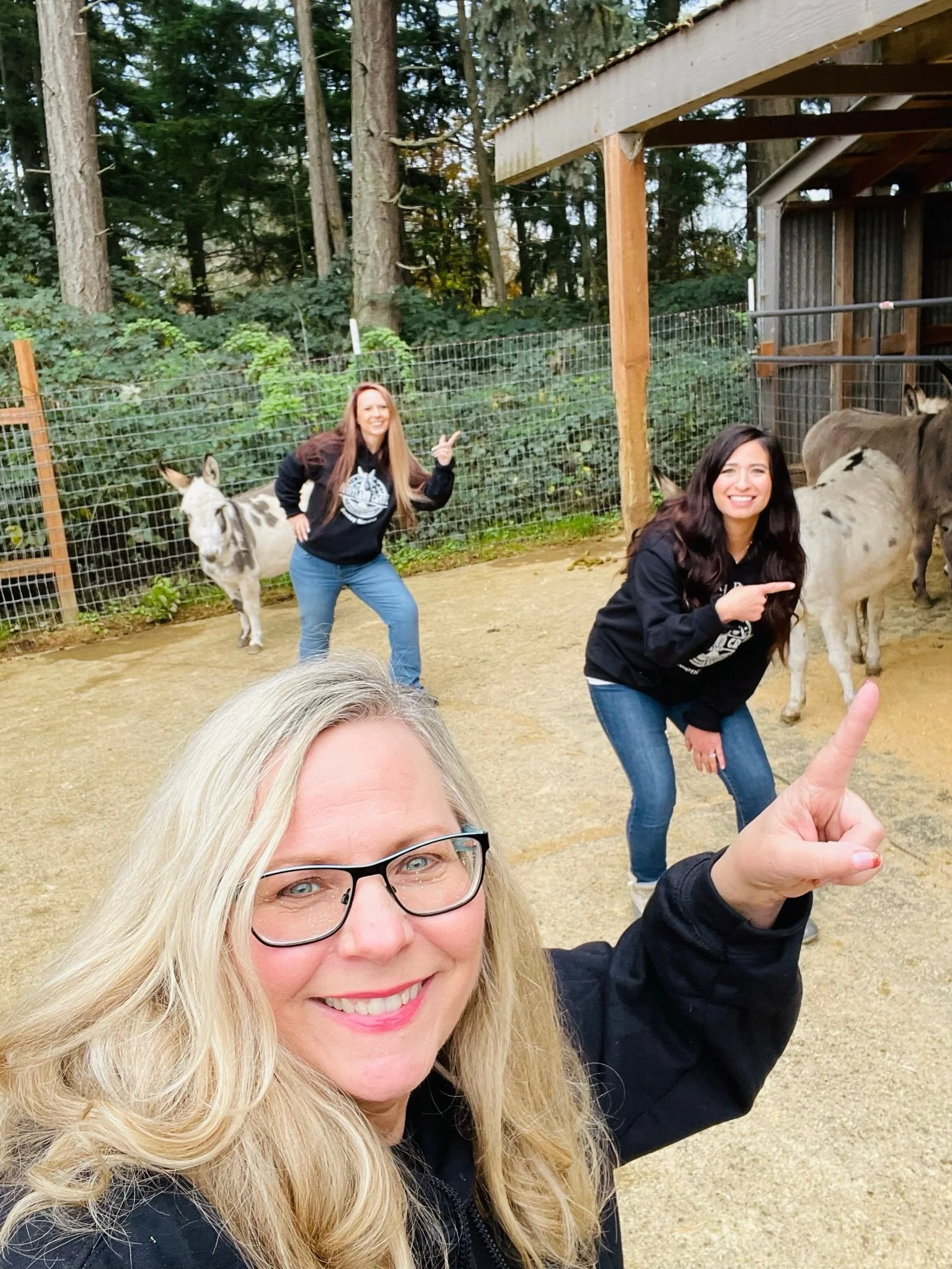 Three women posing happily with donkeys in an outdoor farm enclosure, with trees and fencing in the background.