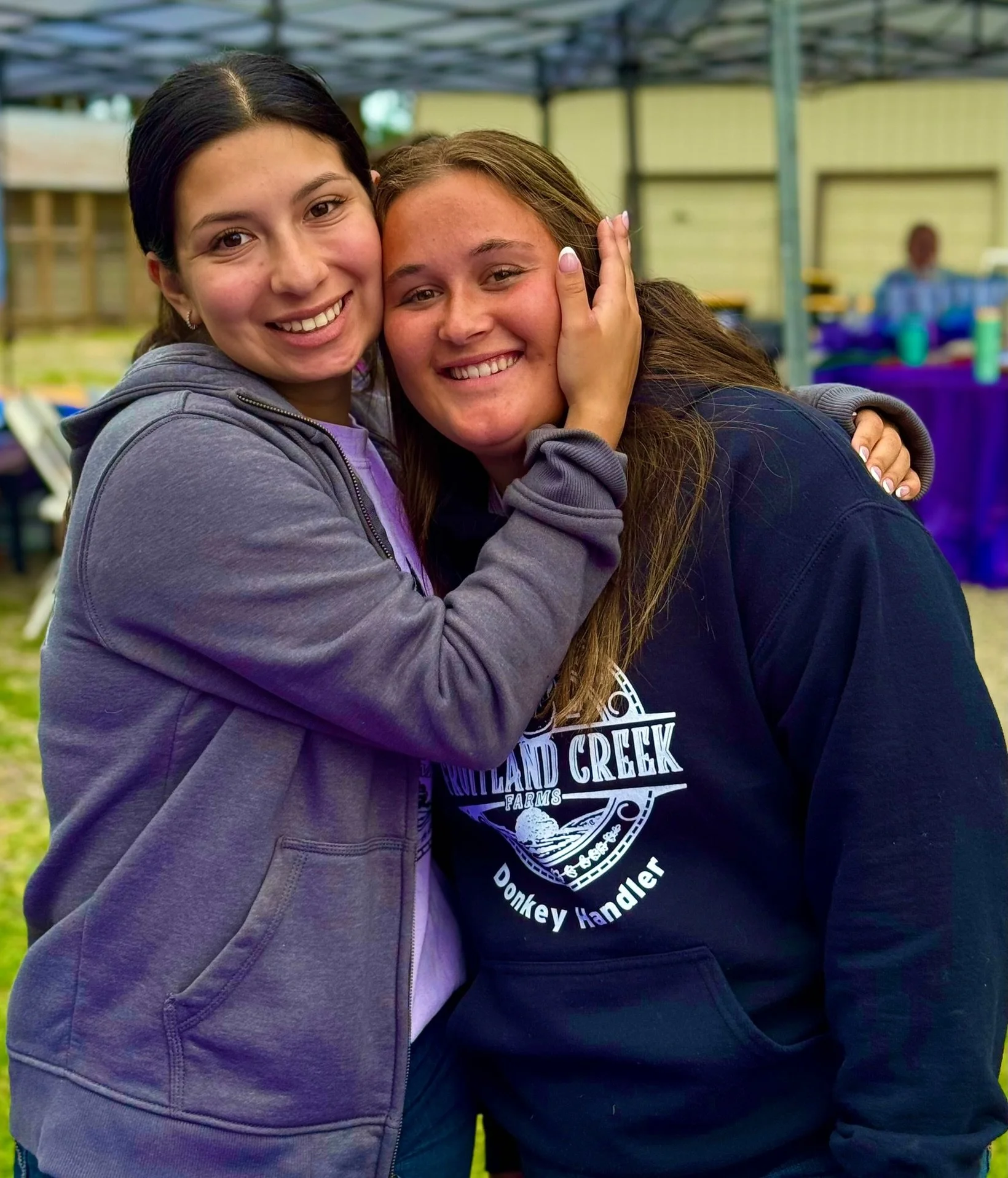 Two young women hugging and smiling at an outdoor gathering.