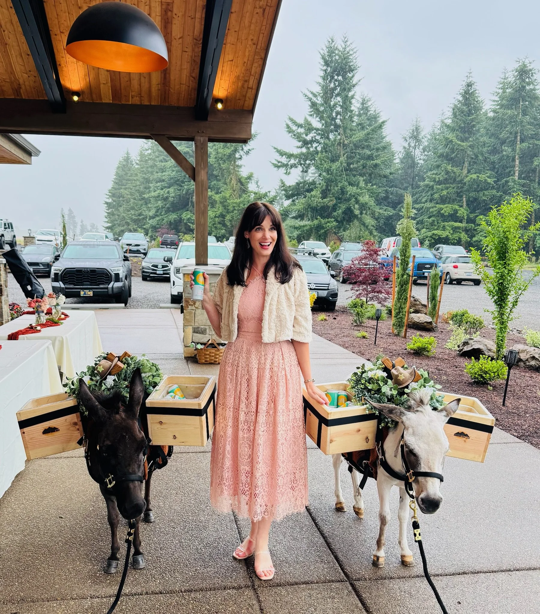 A woman in a pink lace dress and white jacket standing outdoors, holding a soda can, with two donkeys carrying floral baskets, in a parking lot near trees and a wooden shelter.