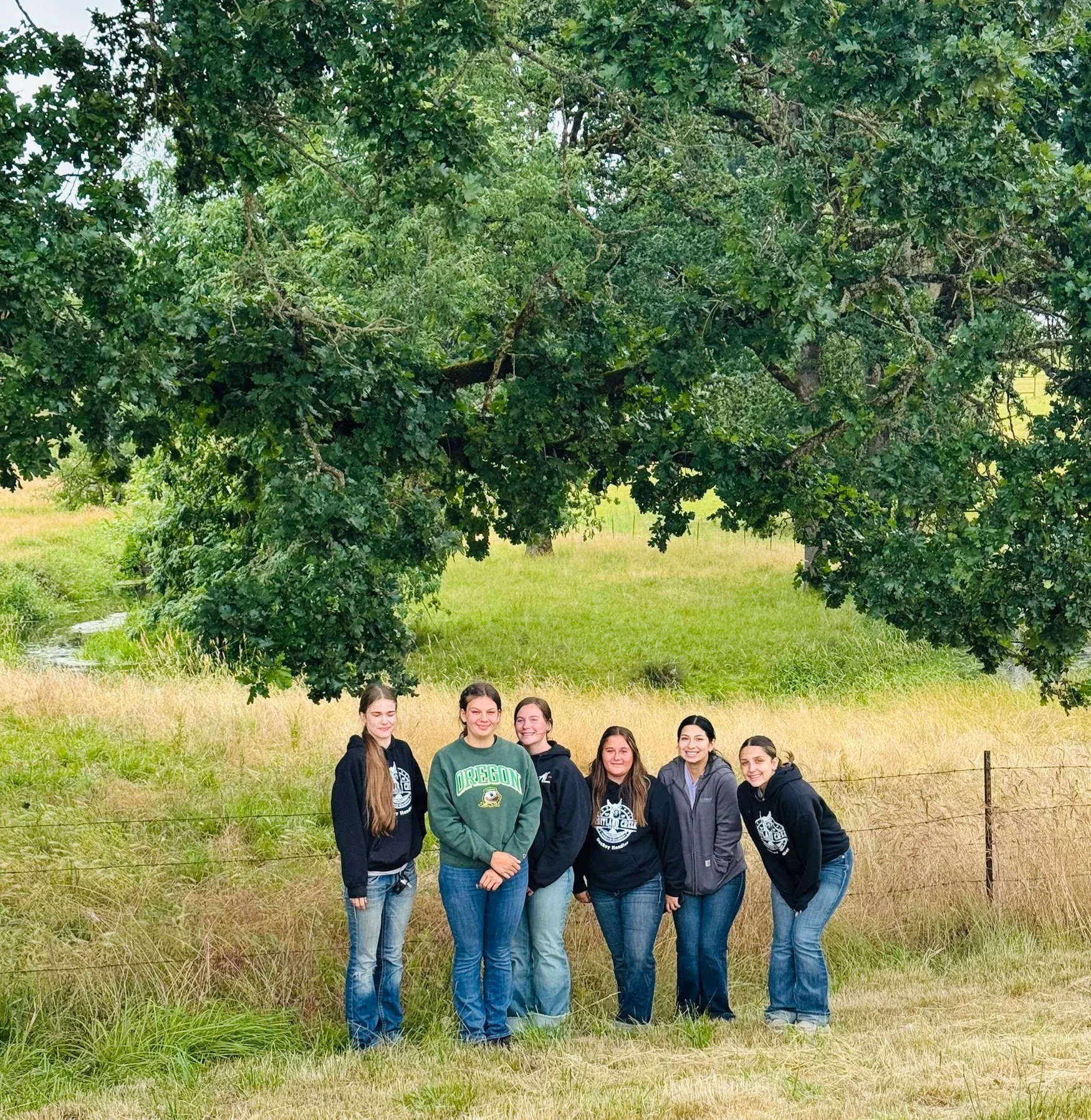 Five young women standing under a large tree in a grassy field, smiling at the camera.