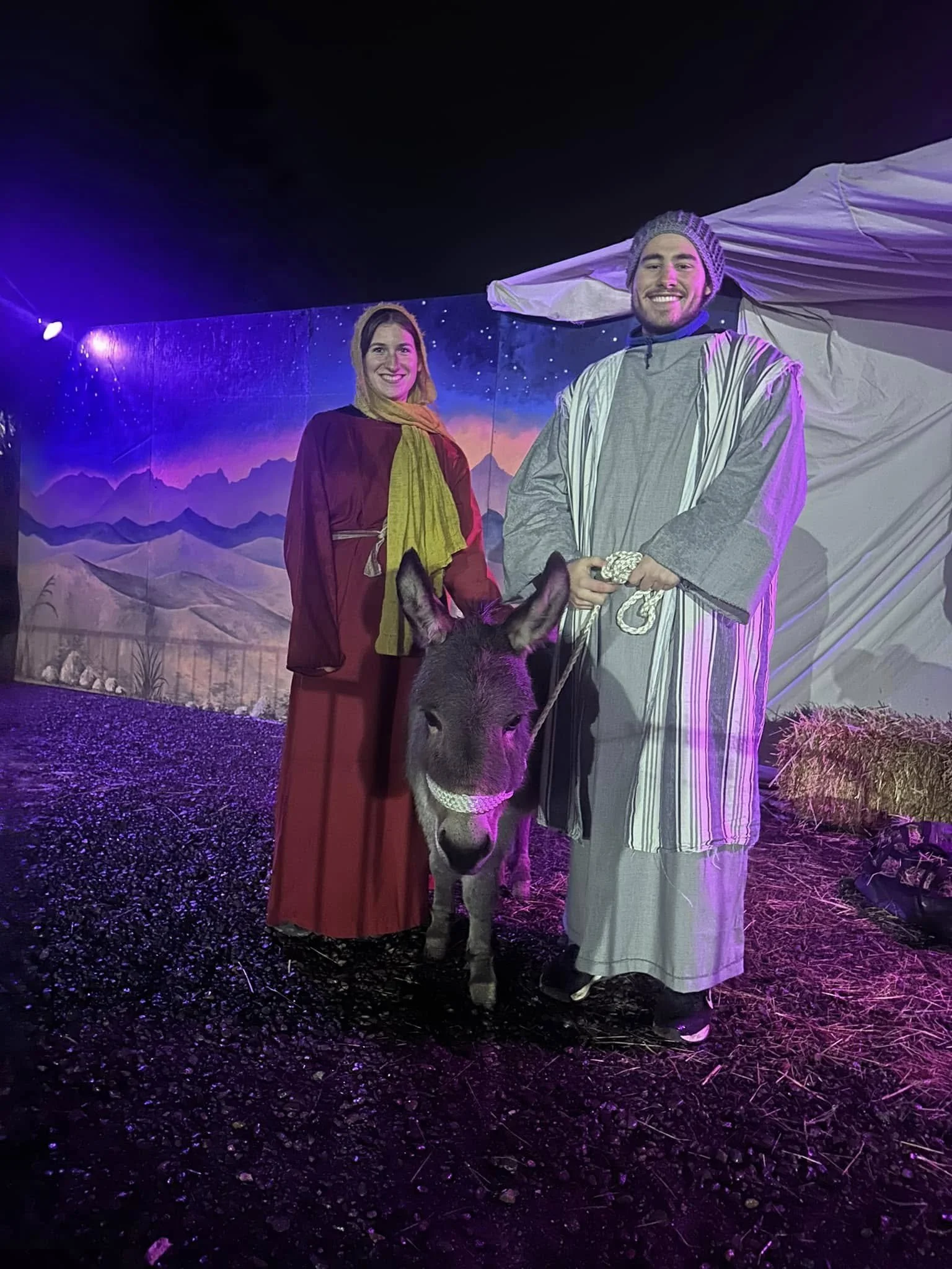 Two people dressed in biblical costumes stand next to a donkey with a painted nativity scene background.