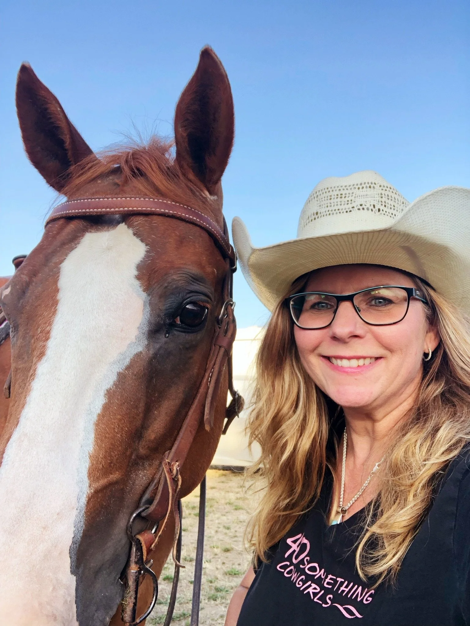 A woman with long blonde hair, wearing glasses and a white cowboy hat, smiling next to a brown and white horse with a white blaze on its face, against a clear blue sky.