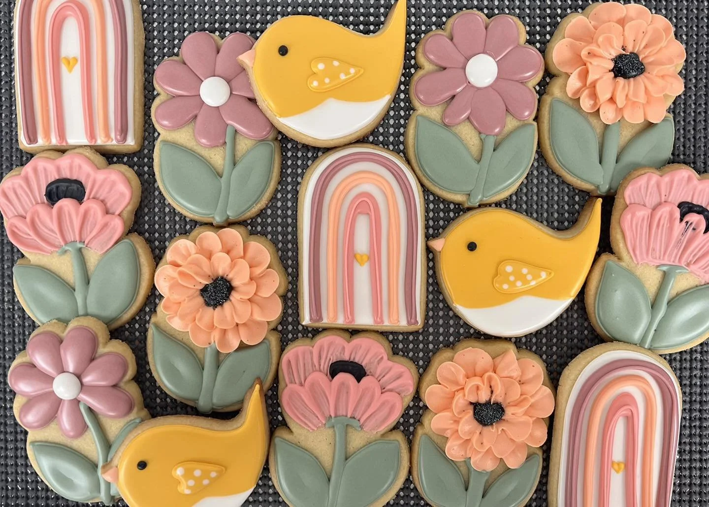 Decorated cookies featuring pink flowers with black centers, orange flowers with black centers, yellow ducks with white and orange details, and rainbow patterns in pastel colors arranged on a black textured surface.