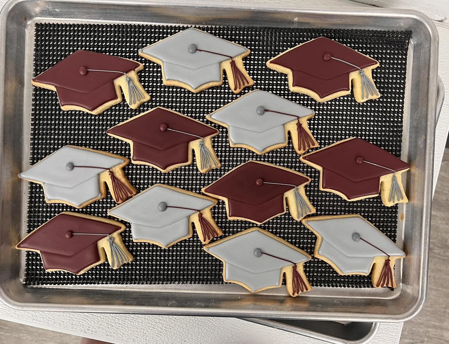Decorated cookies shaped like graduation caps, with some caps in red and others in gray, placed on a black textured mat inside a metal baking sheet.
